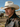Ranch cowboy wearing a wool felt hat with a brown velvet hat band around the crown, western style jacket, standing at a ranch stable under open sky.